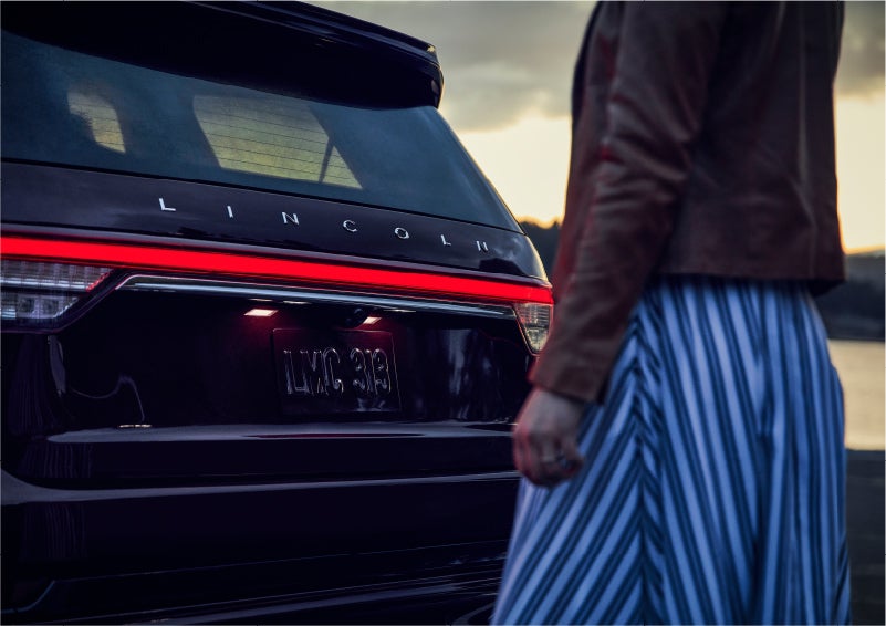 A person is shown near the rear of a 2023 Lincoln Aviator® SUV as the Lincoln Embrace illuminates the rear lights | Golden Circle Lincoln in Jackson TN